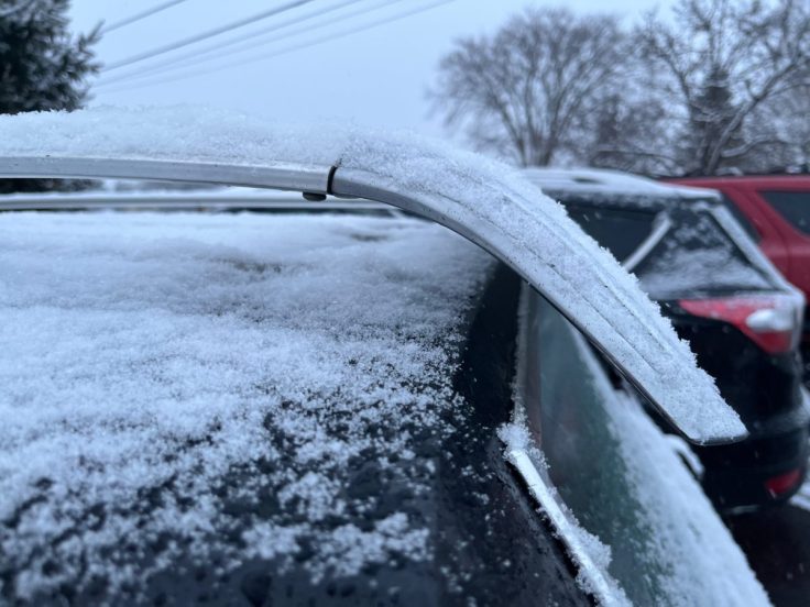 Close-up of a Pontiac Catalina Safari roof covered in a layer of snow, with a metallic roof rack visible, and blurred cars in the background.