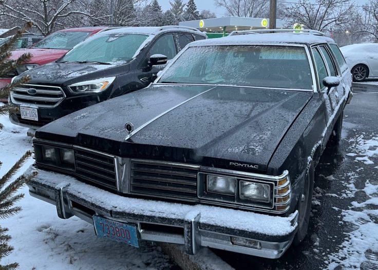 A black Pontiac station wagon covered in snow, parked in a lot with other cars in the background. Snowflakes are visible on the windscreen and hood.