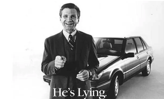 A smiling man in a suit gesturing while standing next to a car, with the text 'He's Lying.' beneath him.