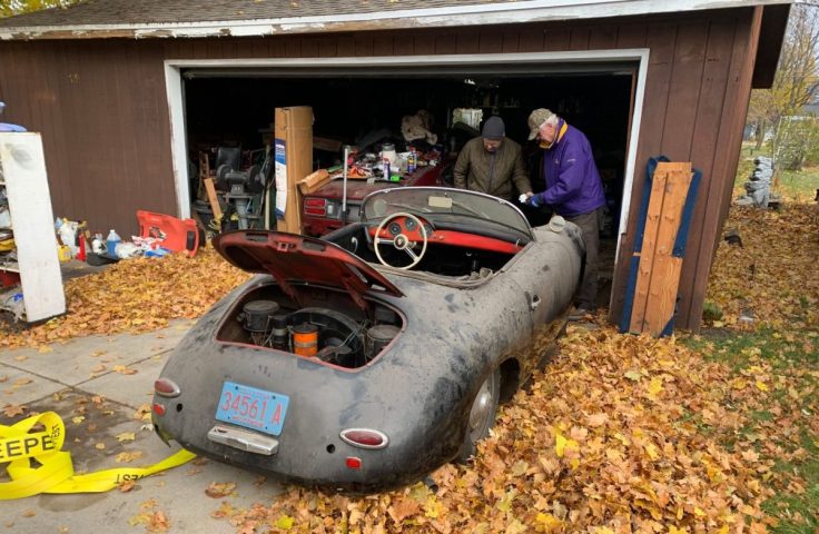 Two men examining a dusty vintage car in a cluttered garage, surrounded by fallen leaves.