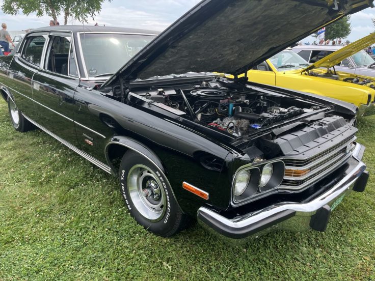 A classic black Ambassador with the hood open, displaying its engine; parked on grass with several other cars in the background.