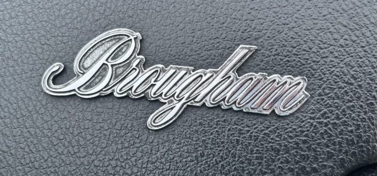 Close-up of a chrome 'Brougham' emblem on a textured black background on 74 AMC Ambassador.