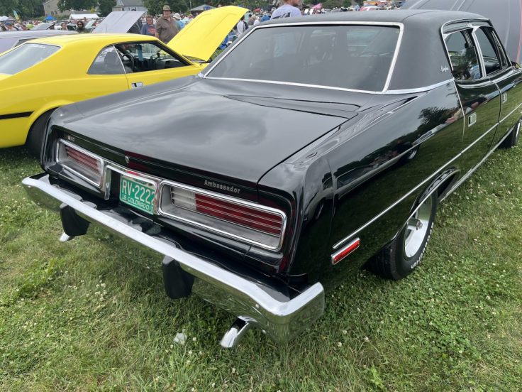 Rear view of a black 74 AMC Ambassador with chrome accents and a distinctive design, parked on grass beside a yellow car.