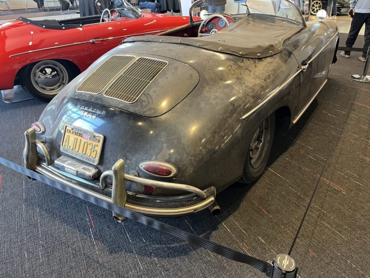 A vintage black Porsche sports car with a dusty exterior, featuring a convertible top and distinctive rear design, alongside a red classic car in the background.