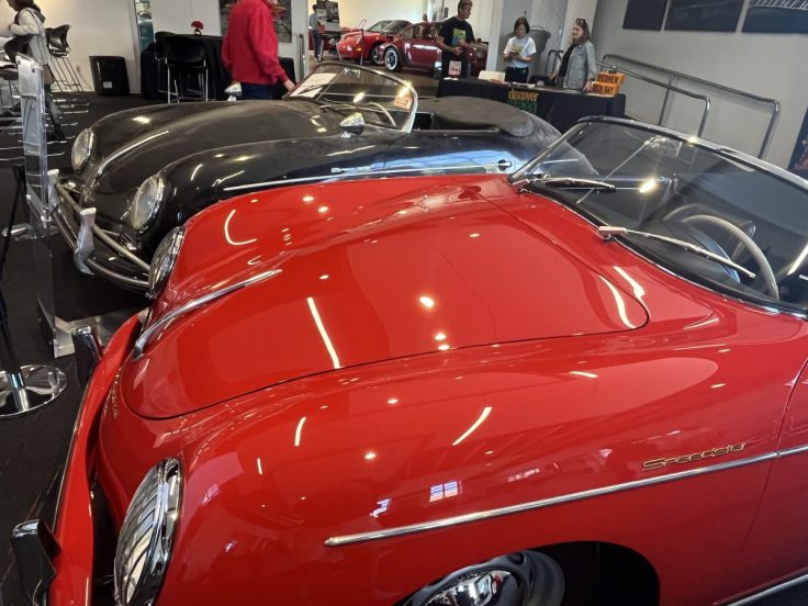 A close-up view of a vibrant red classic car in the foreground, with a black classic car directly behind it. The background features other vintage cars and people observing them in a showroom.